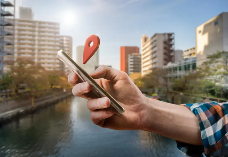 Mano sosteniendo un teléfono inteligente con un icono de ubicación rojo flotando sobre un entorno urbano con edificios