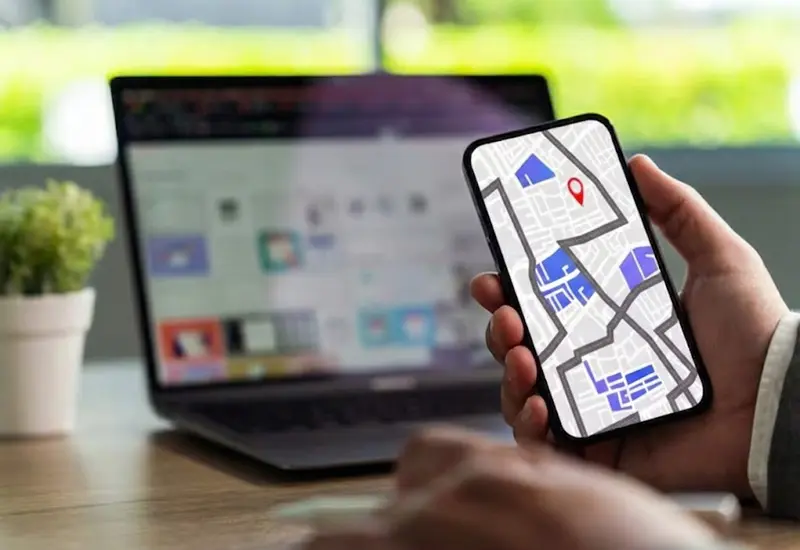 Persona consultando un mapa digital con un pin rojo en su smartphone frente a un ordenador en una oficina.