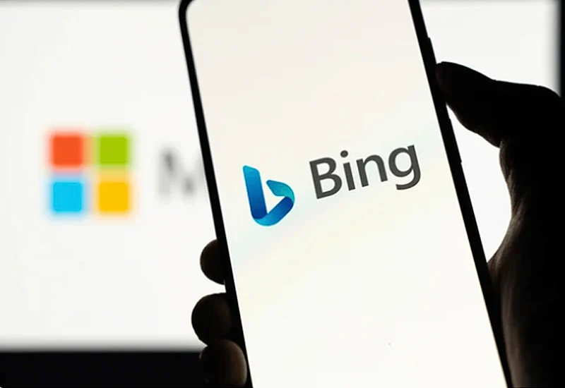 Mano sosteniendo un smartphone con el logo de Bing en pantalla y el logo de Microsoft desenfocado en el fondo